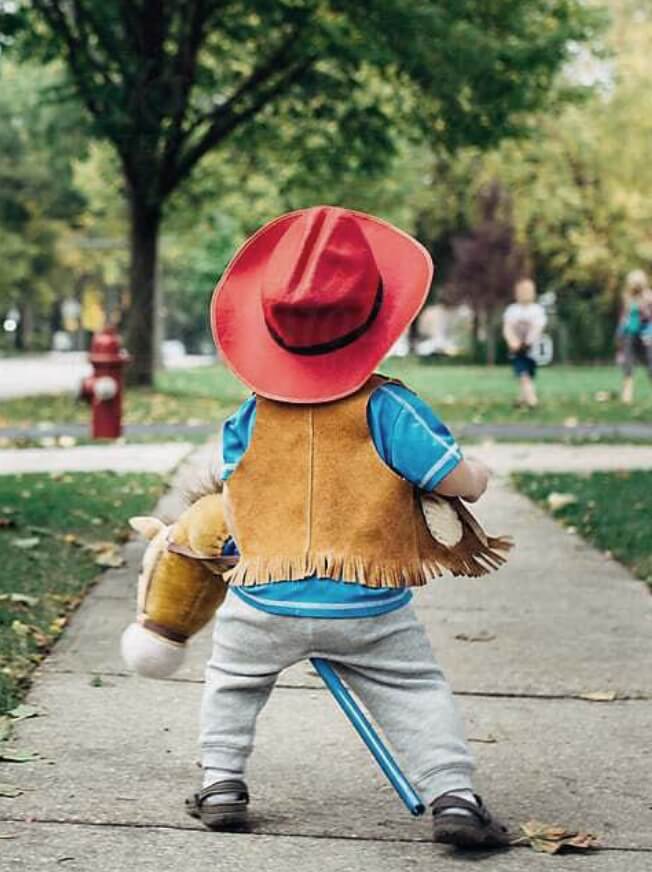A toddler dressed as a cowboy walking down a sidewalk in a family-friendly neighborhood.