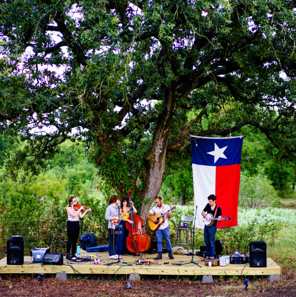 A folk band performs outdoors next to a large Texas flag, near new homes in Goodland TX.
