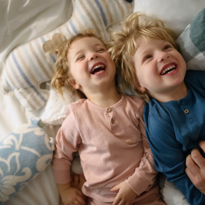 Two young children laughing while lying side by side on a bed in their new home in Mansfield, TX.