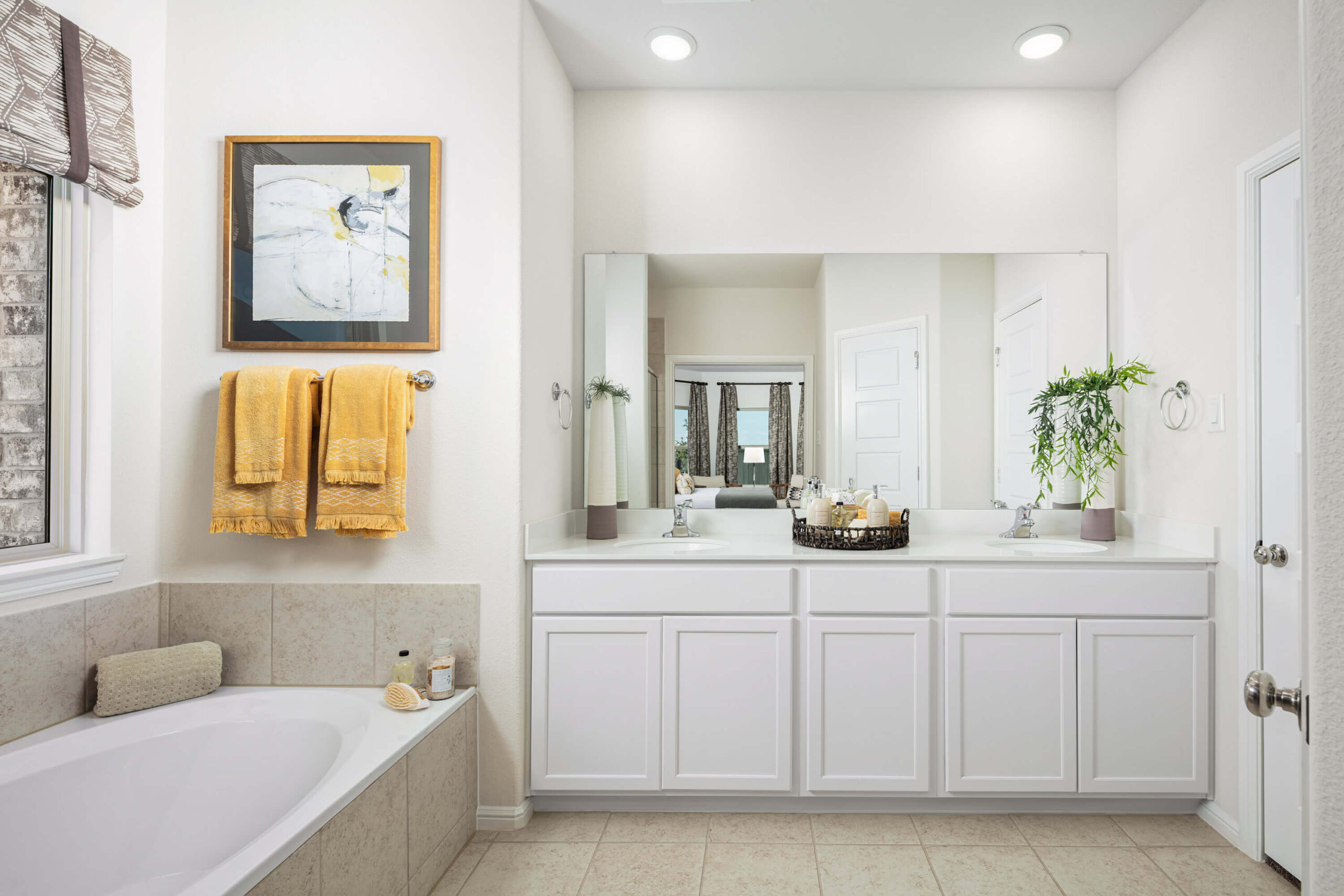 A bright and clean bathroom in new homes in Goodland TX with dual vanity sinks, a large mirror, and a bathtub, accented with yellow towels and green plants.