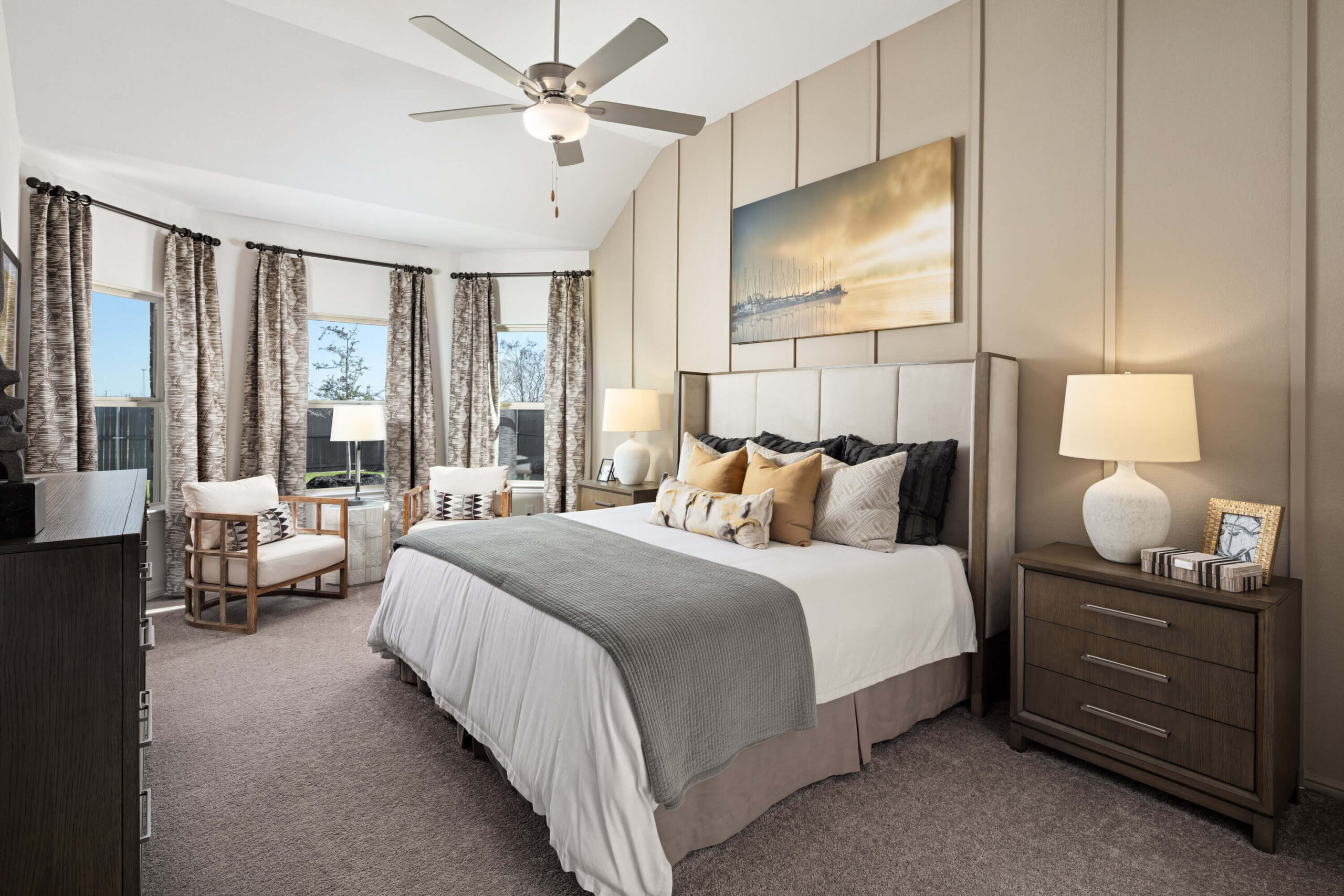 A well-appointed bedroom in new home communities in Mansfield TX, featuring a queen-sized bed with decorative pillows, two nightstands, a ceiling fan, and large windows with patterned curtains.