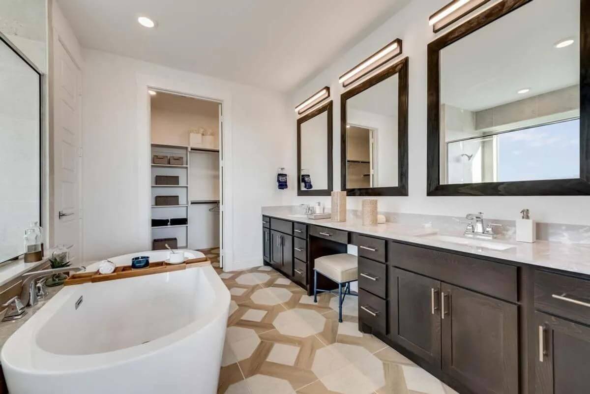 Modern bathroom interior in new homes in Goodland TX with dual vanity sinks, large mirrors, a freestanding bathtub, and geometric tiled flooring.