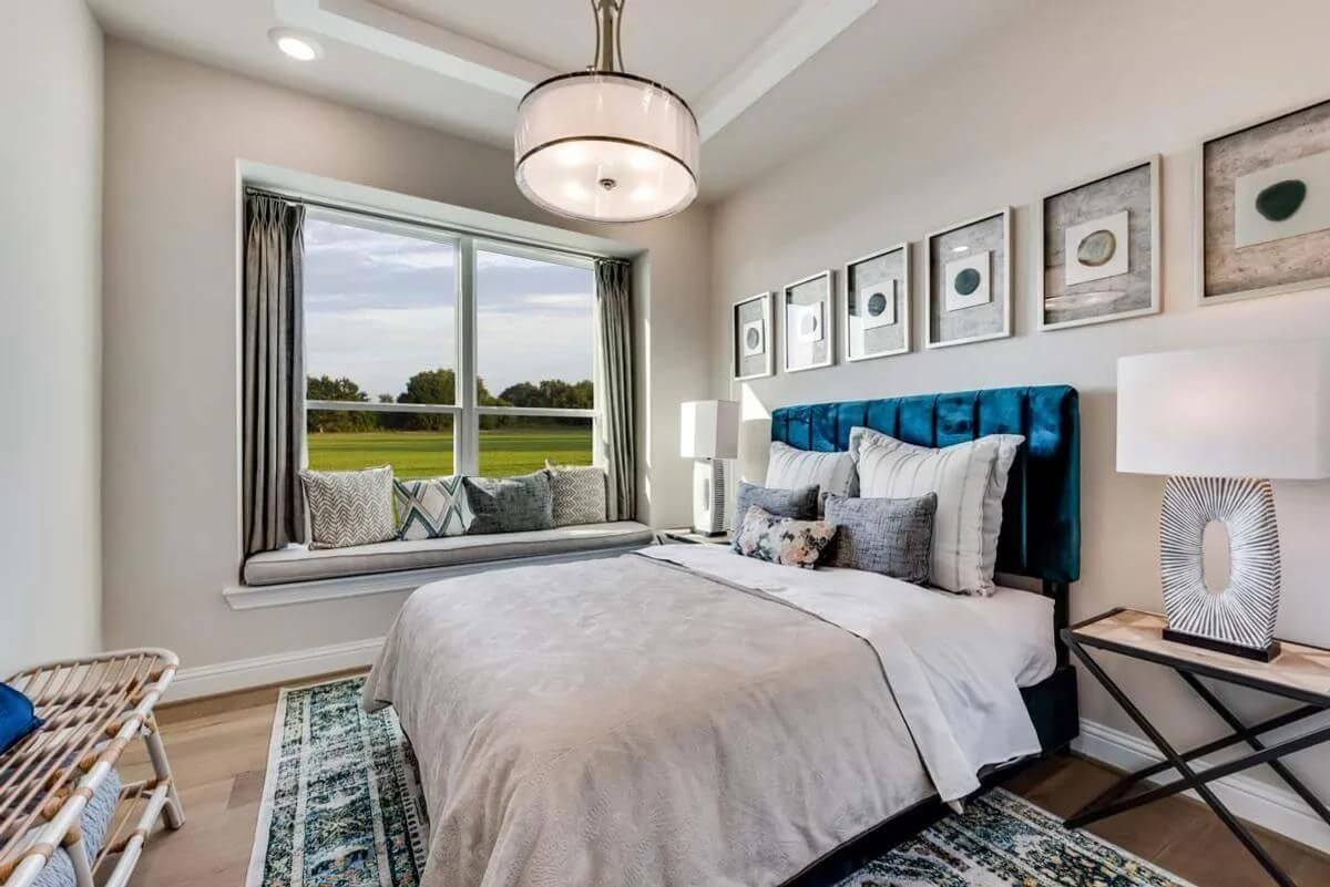 A well-lit, modern bedroom in new homes in DFW with a blue upholstered headboard, matching pillows, wall art, and a view of the outdoors.
