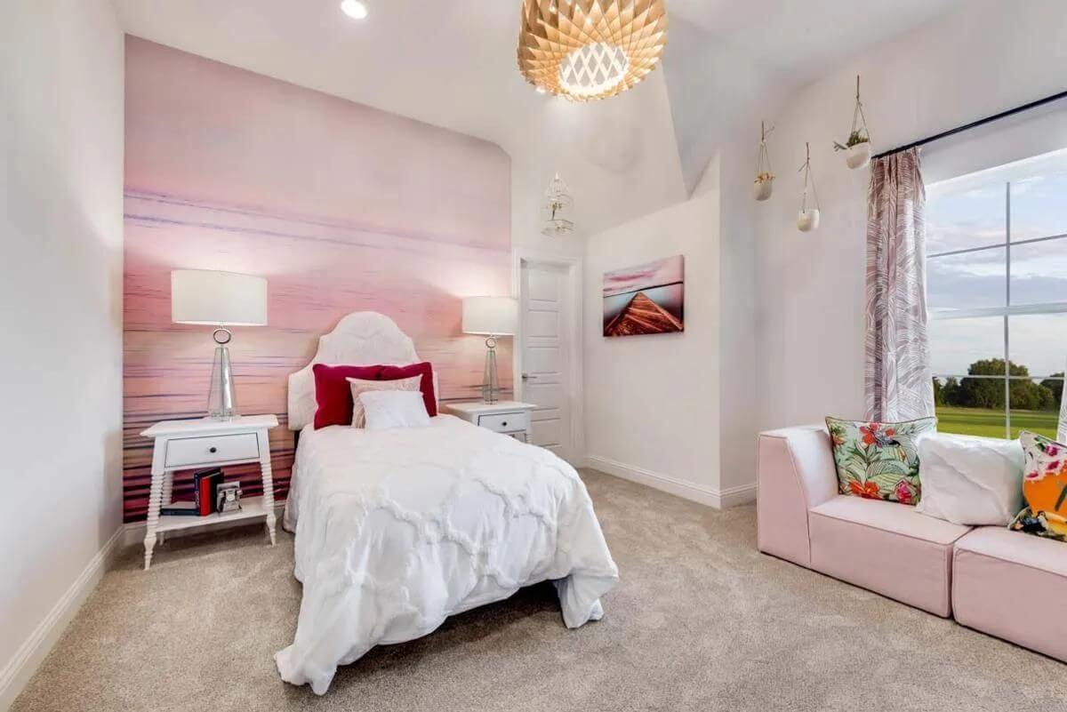 A well-lit, contemporary bedroom in a new home community in Mansfield, TX, with a white bed, pink accents, and a chaise lounge by the window.
