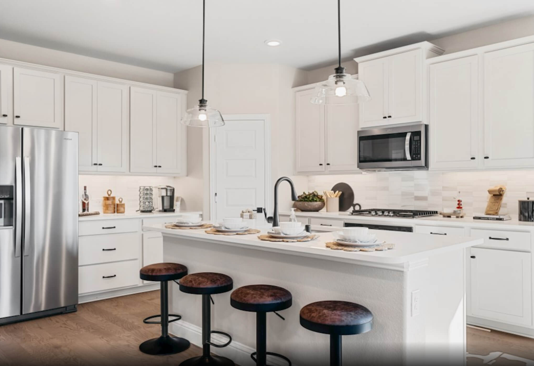 A modern kitchen in new homes in Goodland, TX, with white cabinetry, stainless steel appliances, and a breakfast bar with stools.