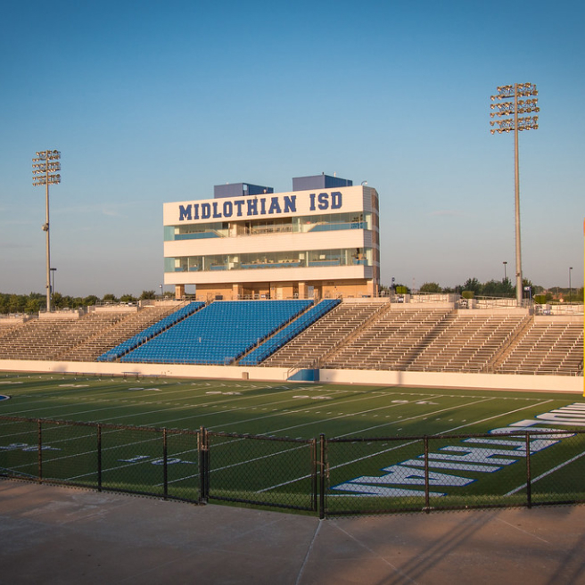 A high school football stadium with blue and white bleachers under a clear sky at dusk, near new homes in Goodland TX.