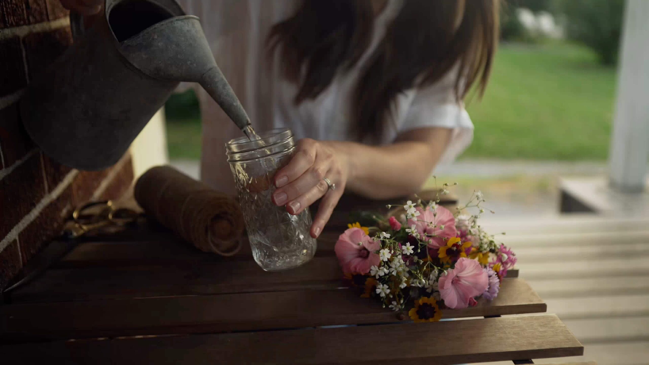 Woman pouring water from a metal watering can into a glass jar beside a bouquet of wildflowers on a wooden bench, evoking the serene lifestyle available in new home communities in Mansfield TX.