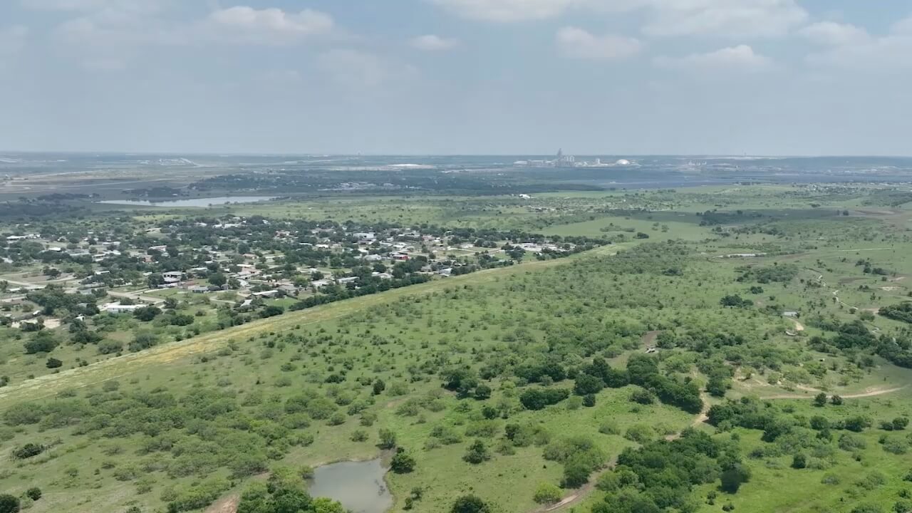 Aerial view of a lush green landscape with scattered trees, a new home in Mansfield TX, and industrial structures in the distance.