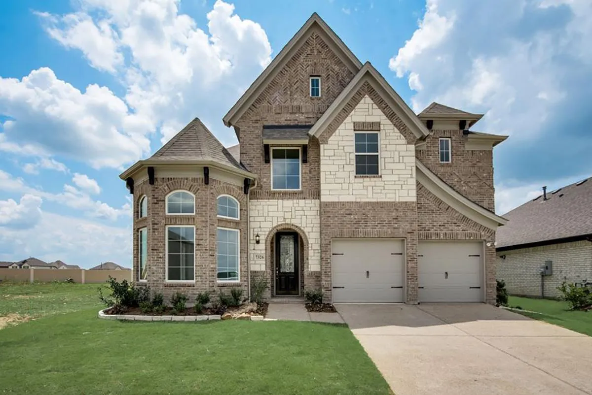 Two-story brick house with arched entryway and double garage doors in a new home community in Mansfield, TX, under a clear sky.
