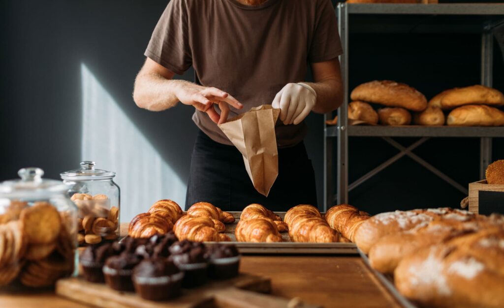 A baker packaging croissants in a paper bag at a bakery display counter in Mansfield, TX.
