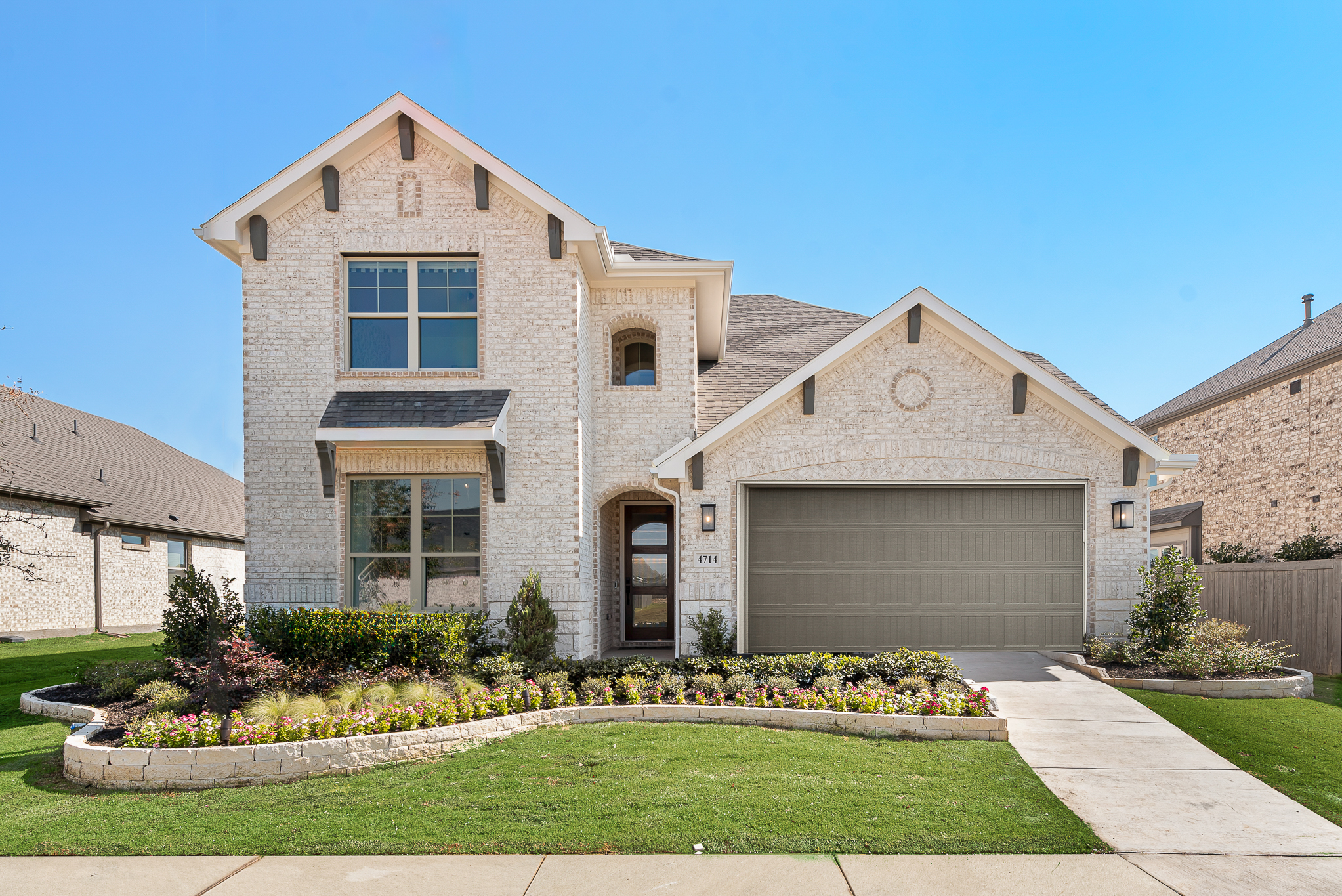 A two-story brick house with a manicured lawn, flower beds, and a gray garage door, set against a clear blue sky.