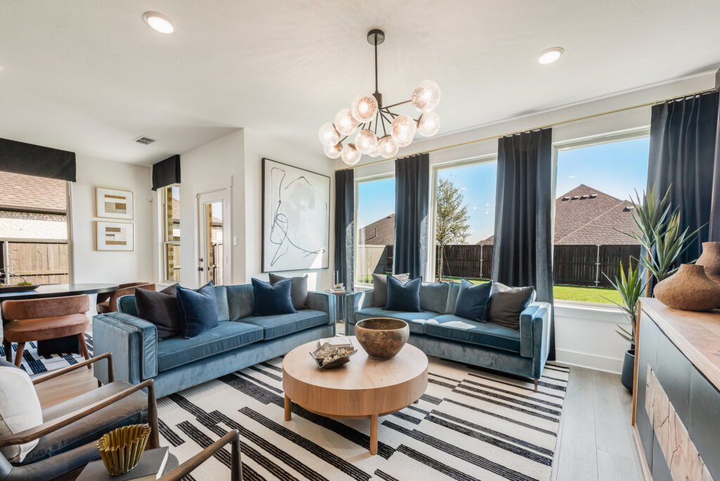 Modern living room with two blue sofas, a round wooden coffee table, and a geometric chandelier. Large windows with black curtains overlook a backyard.