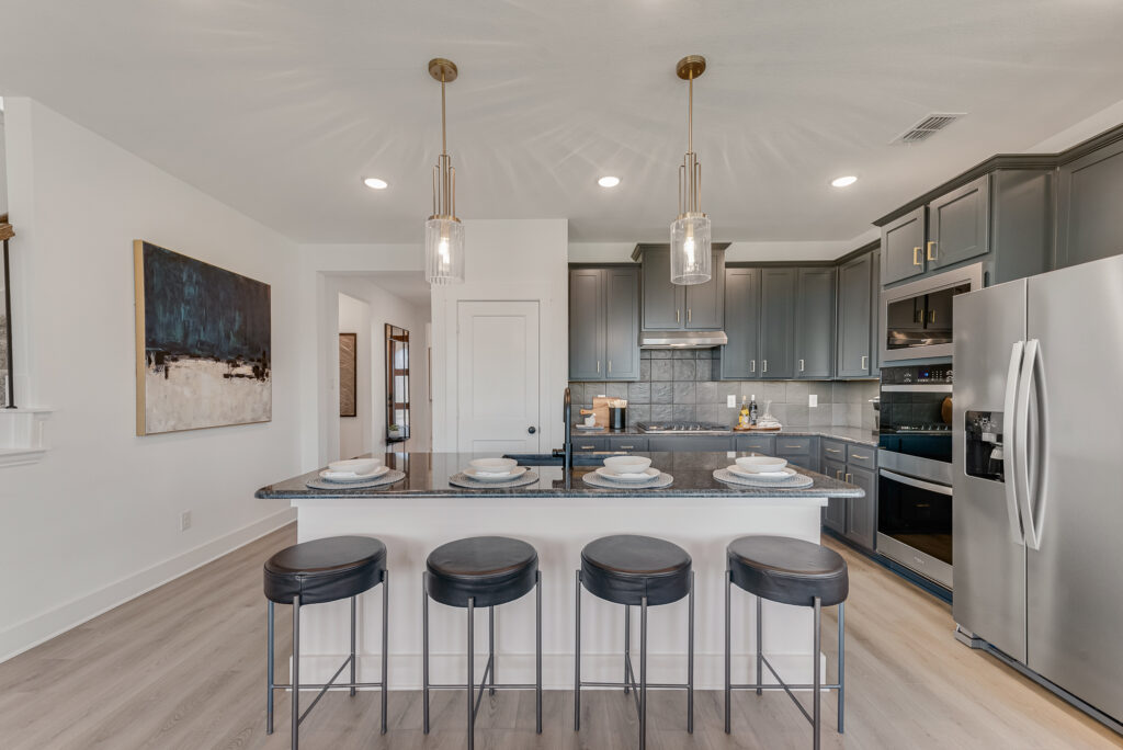 Modern kitchen with an island, bar stools, and stainless steel appliances. Gray cabinets, pendant lights, hardwood floor, and a large painting on the wall.