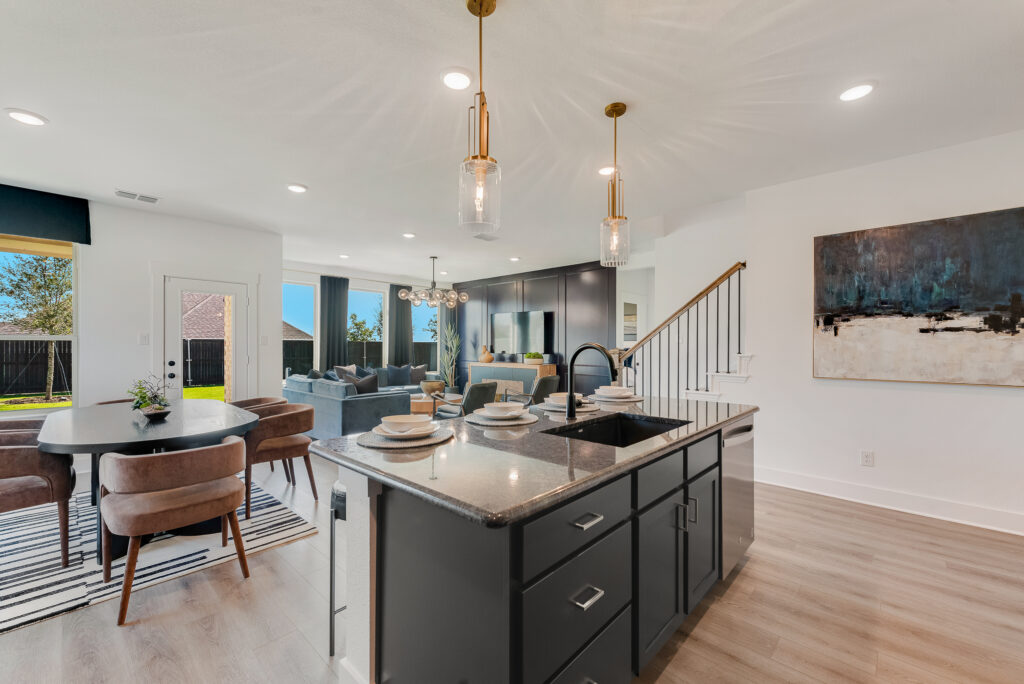 Modern open-concept kitchen and living area with gray cabinets, island, dining table, and seating area. Large windows and pendant lights enhance the bright space.