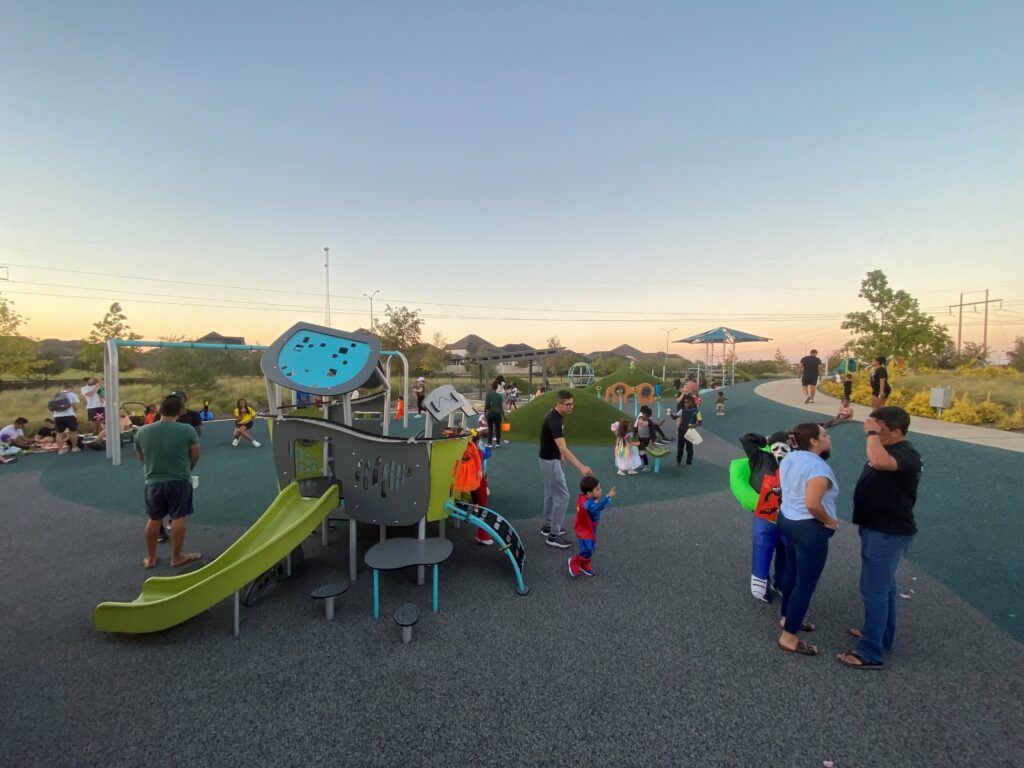 People enjoy a playground at sunset with children on play structures and adults standing nearby.