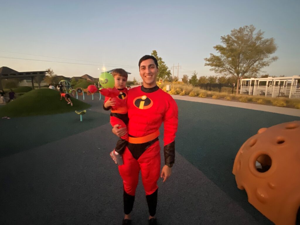 An adult and a child wearing superhero costumes stand together in an outdoor park setting at dusk.