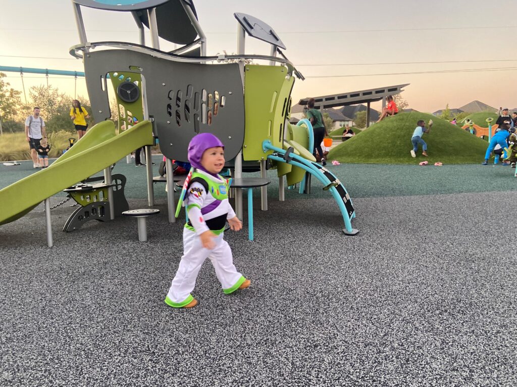 Child in a Buzz Lightyear costume walks in a playground area with various play structures and other children in the background.