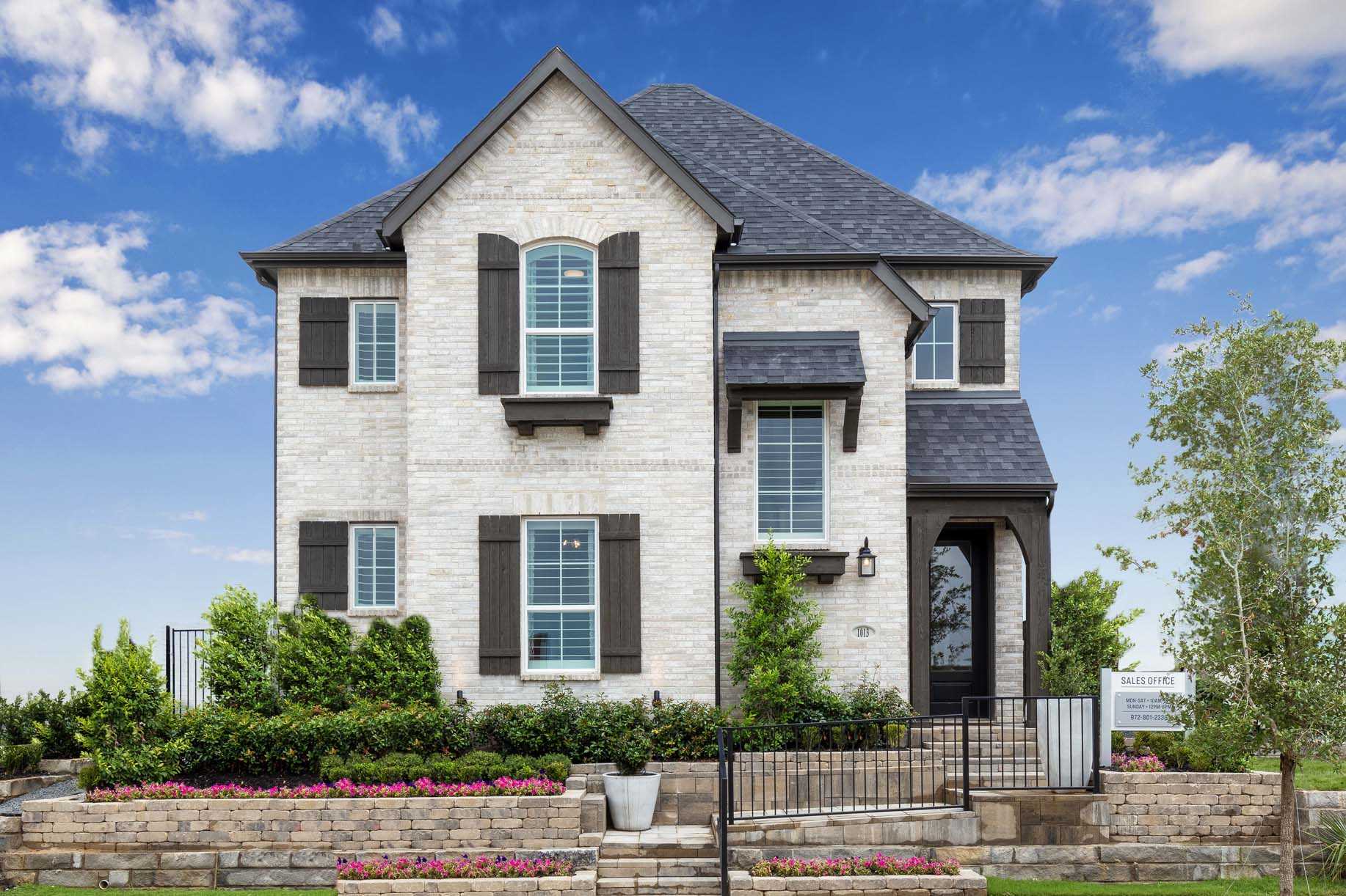 Two-story brick house with dark shutters, a gabled roof, landscaped front yard, flower beds, and a small staircase leading to the front door under a blue sky.