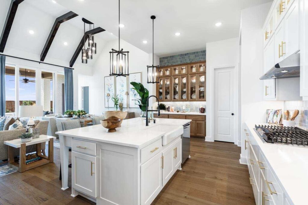 Modern open-concept kitchen with white cabinets, a large island, wood flooring, and pendant lights, adjacent to a living area with large windows and exposed ceiling beams.