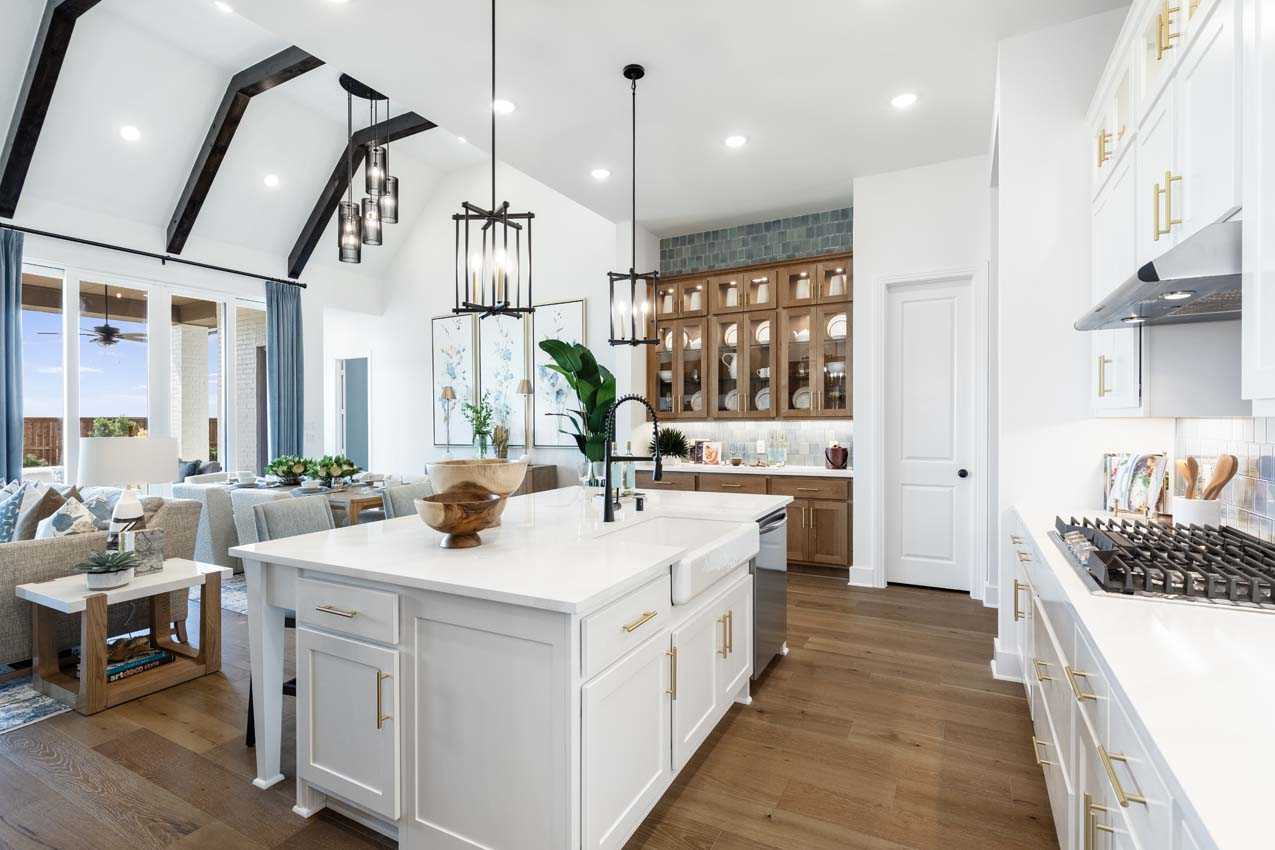 Modern open-concept kitchen with white cabinets, a large island, wood flooring, and pendant lights, adjacent to a living area with large windows and exposed ceiling beams.