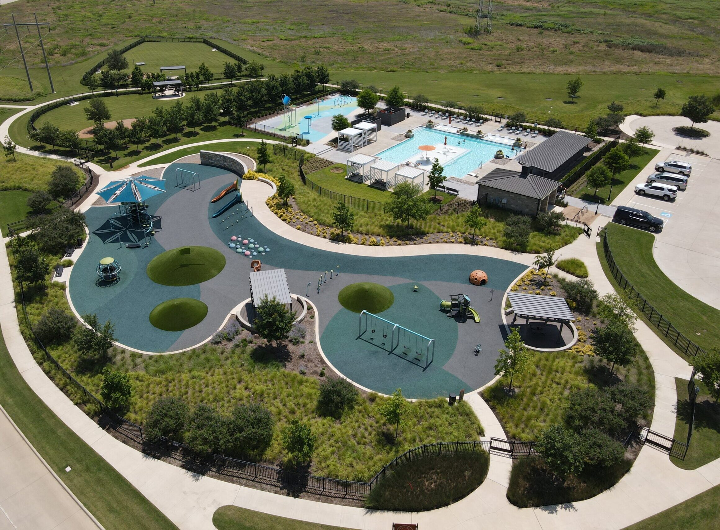 Aerial view of a public park in communities with pools near me, featuring a playground, green spaces, walking paths, a swimming pool, cabanas, and parking lot on a sunny day.