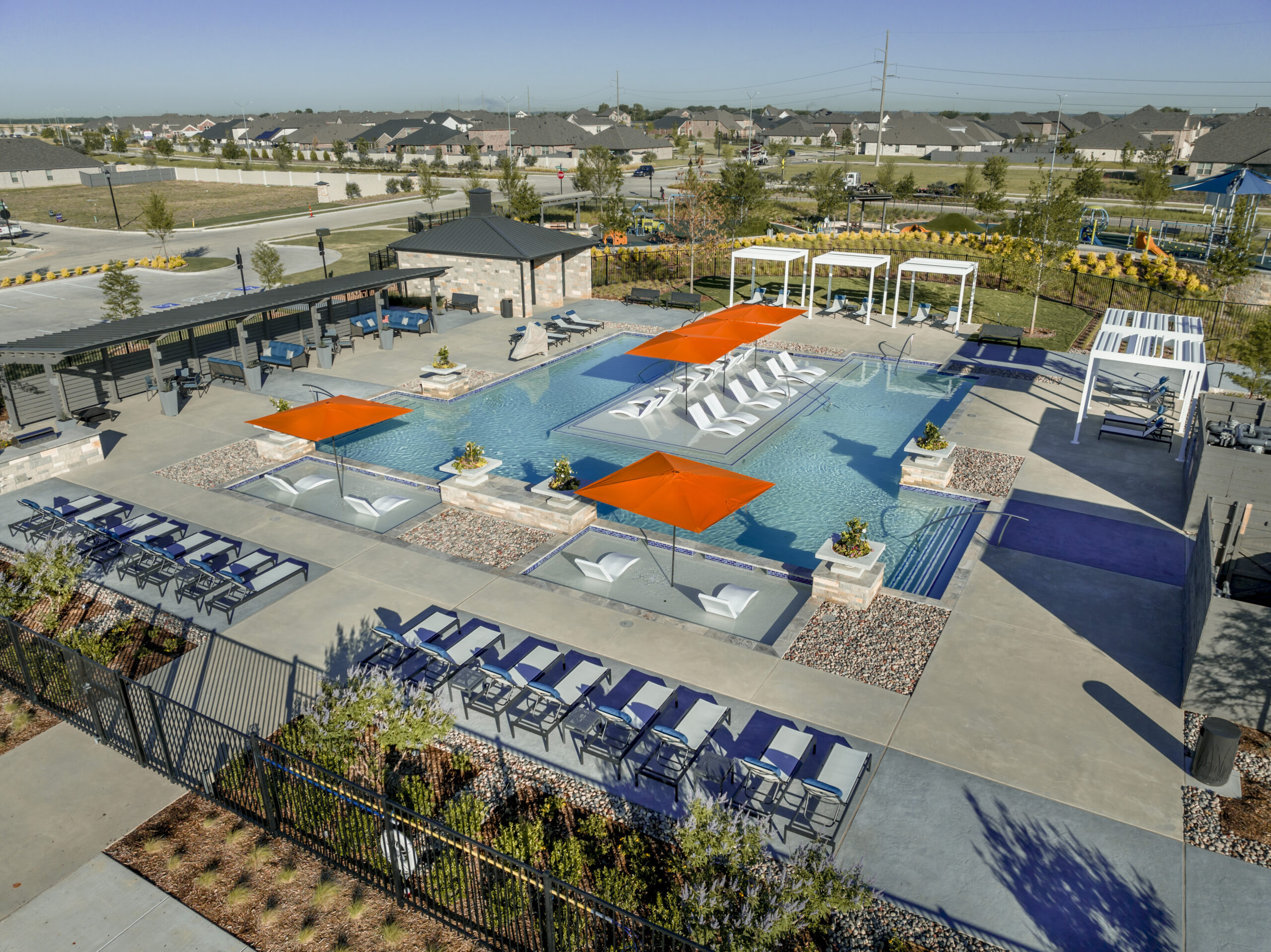 Aerial view of a modern outdoor swimming pool with lounge chairs, cabanas, shaded areas, and landscaped surroundings in a residential neighborhood.