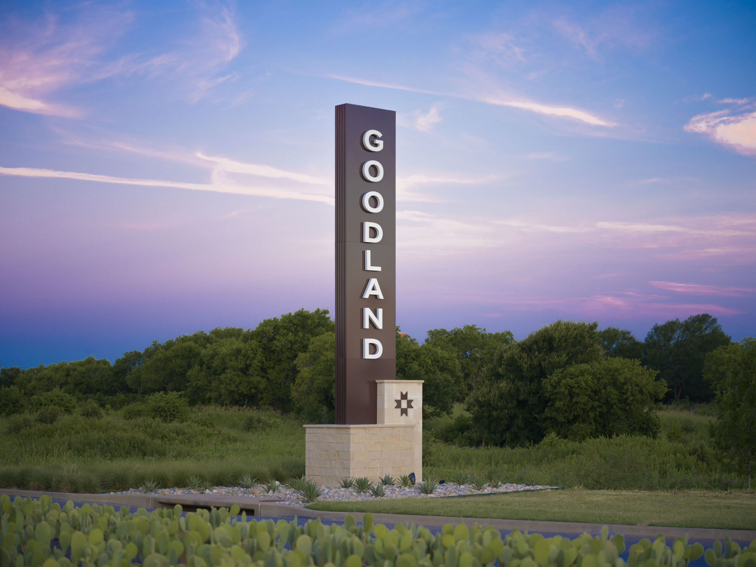 A tall vertical sign reading "GOODLAND" stands on a stone base with greenery and trees in the background at sunset.