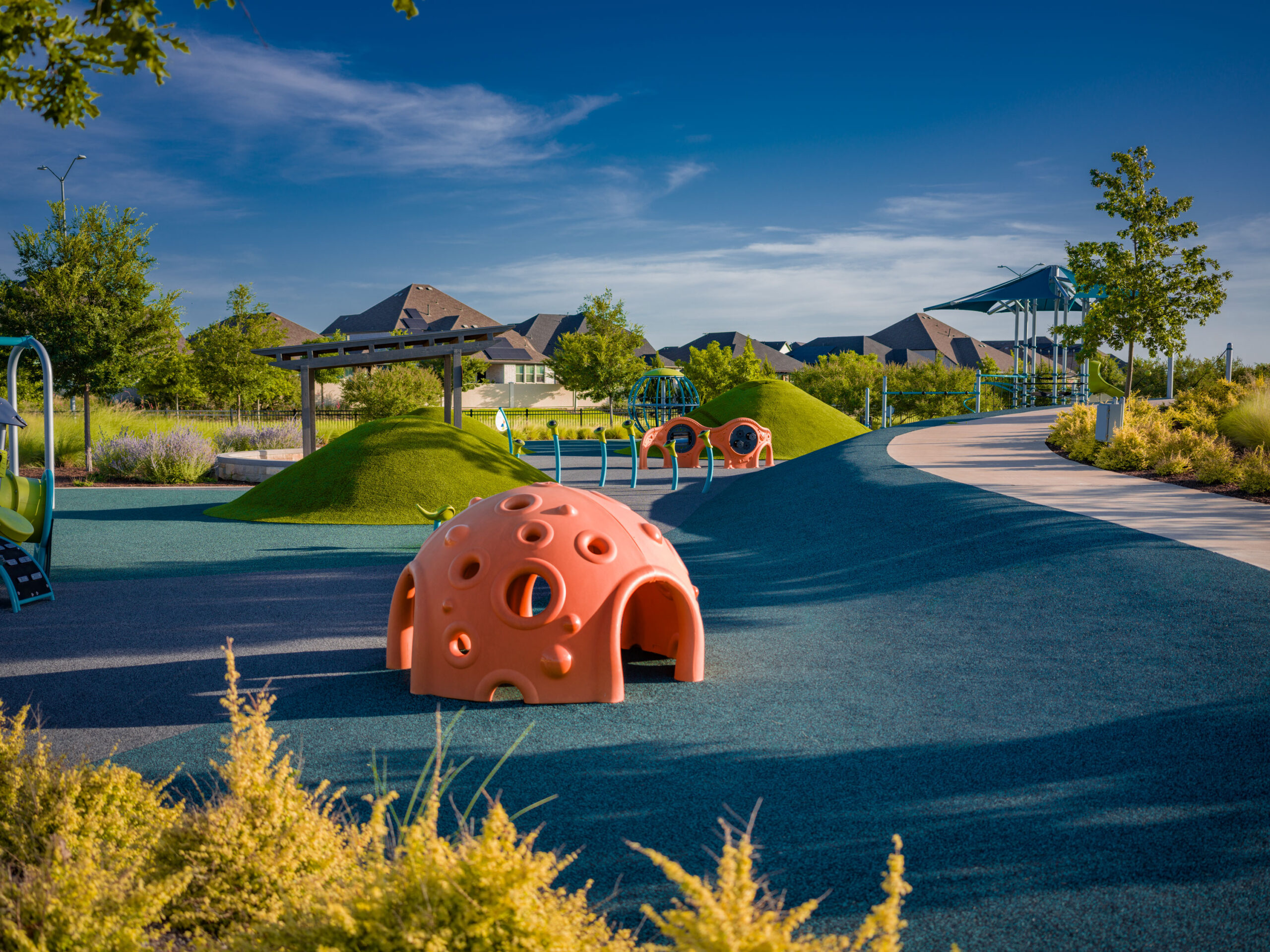 A modern playground with rubber flooring, small green hills, orange climbing domes, play structures, trees, and nearby houses in communities with pools near me under a blue sky.