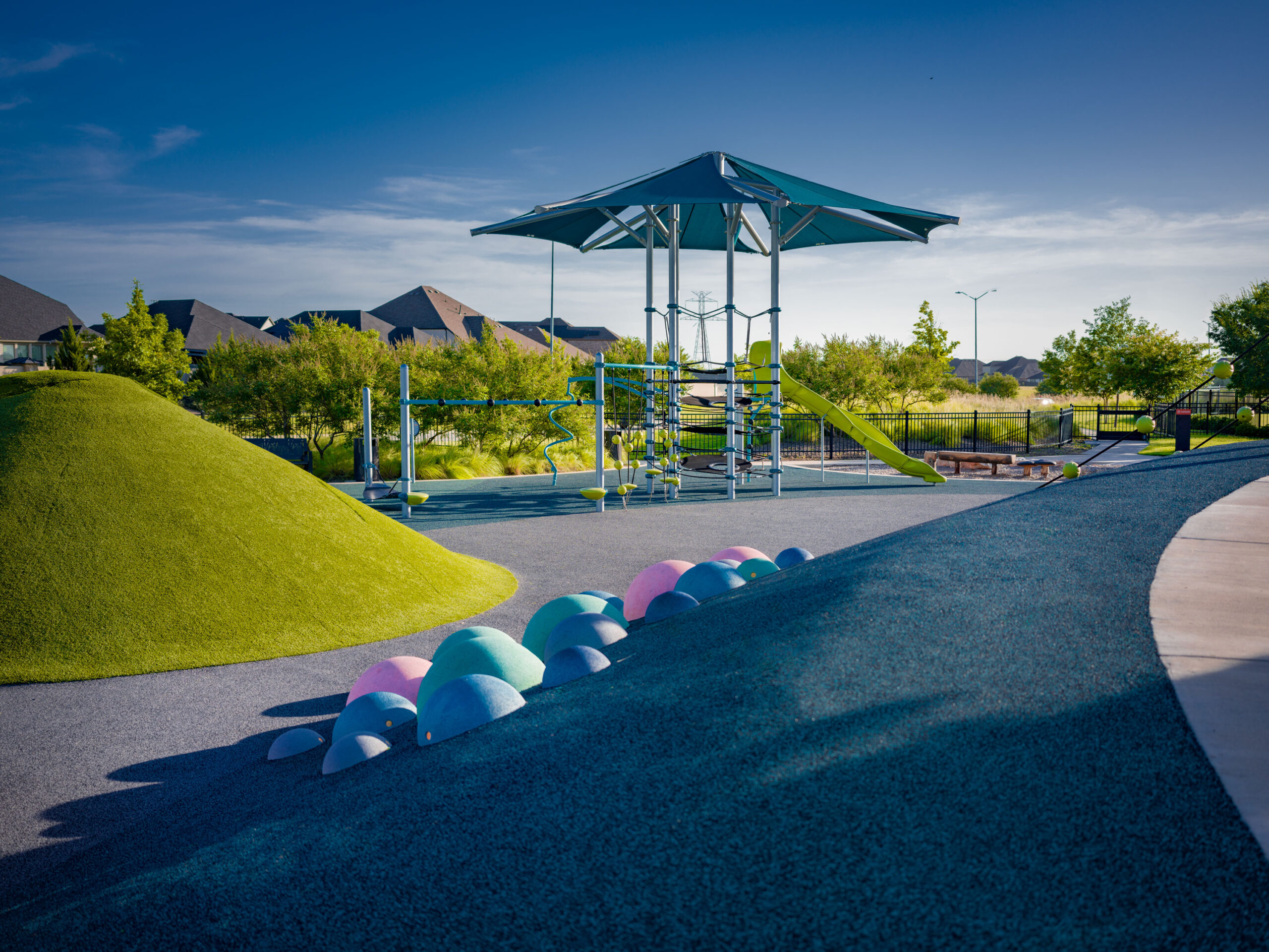 A modern playground with a climbing structure, slide, colorful domes, and grassy mounds under a blue sky—perfect for families searching for communities with pools near me. Houses and trees frame the fun-filled scene.