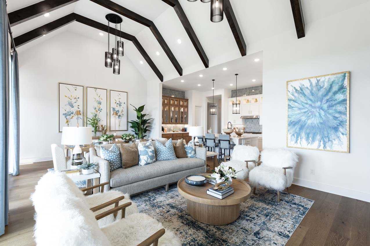 A spacious, modern living room with exposed beams, neutral furniture, blue accents, abstract art, and an open view of the kitchen and dining area.
