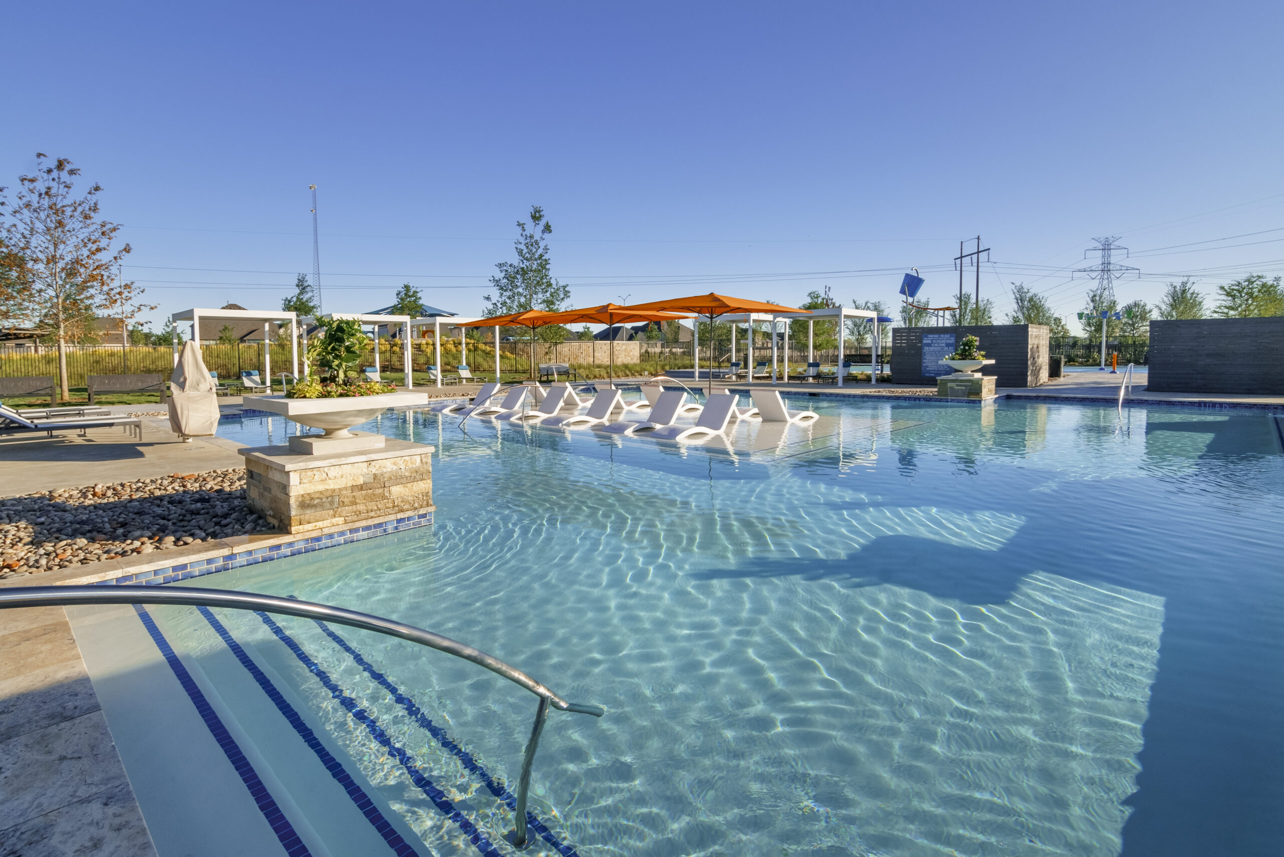 Outdoor swimming pool with submerged lounge chairs, shaded cabanas, and clear blue water on a sunny day—perfect for those searching for communities with pools near me. Landscaped area and utility poles in the background.
