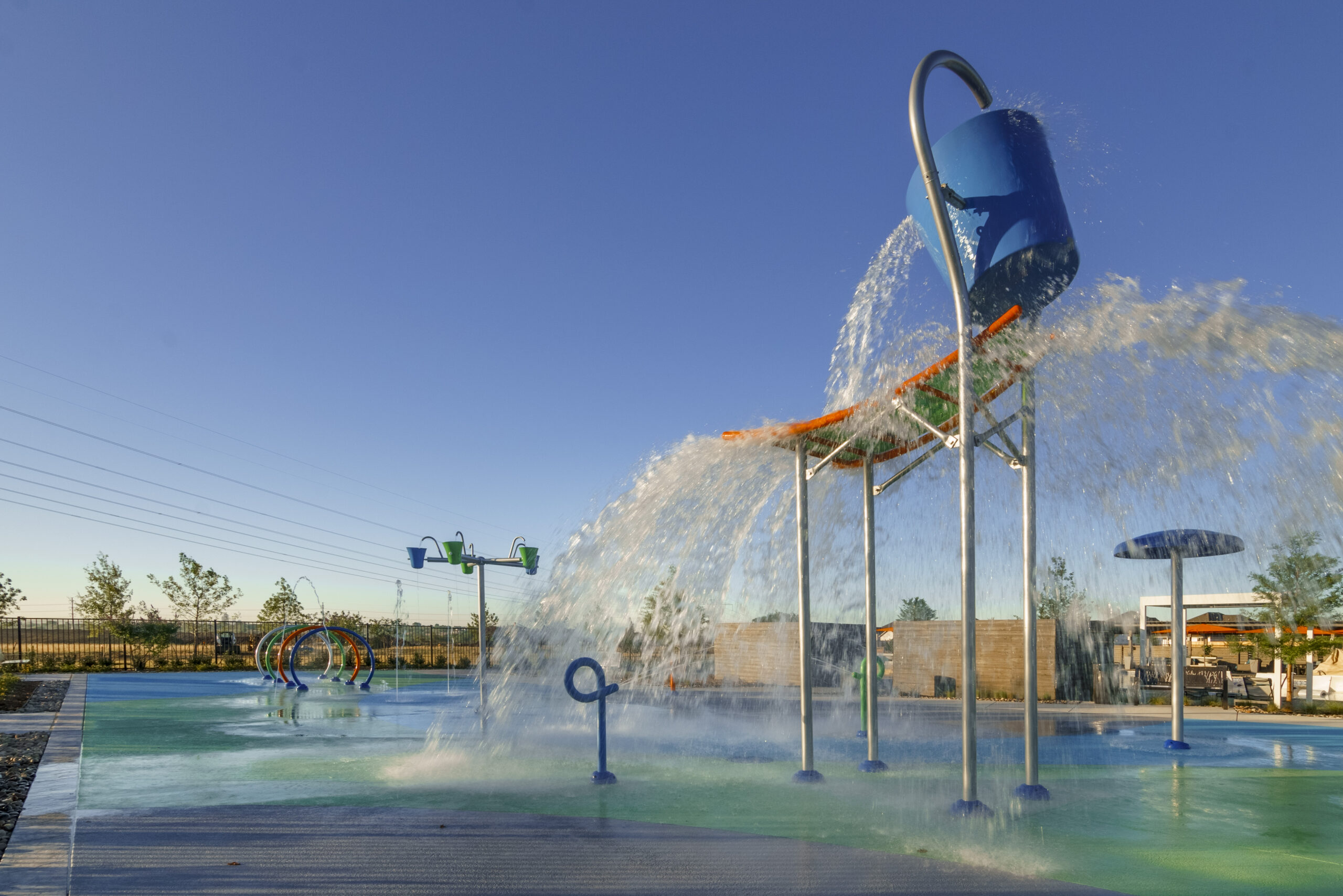 A splash pad with a large tipping bucket pours water onto the play area, surrounded by fun water features and open sky—a perfect spot for families searching for communities with pools near me.