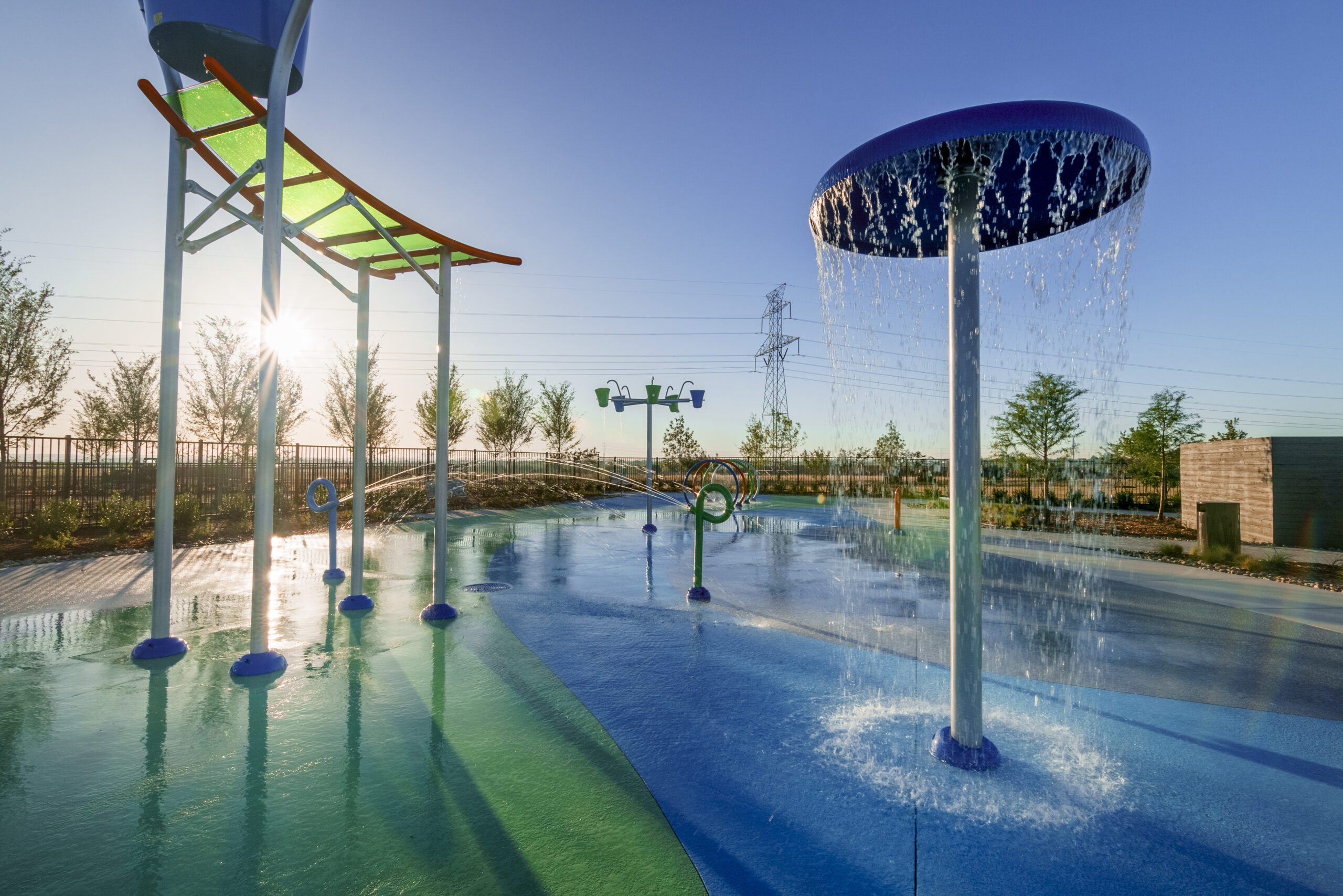Colorful splash pad with water features spraying and pouring water, surrounded by trees and fencing, under a clear blue sky at sunrise—perfect for families seeking communities with pools near me.