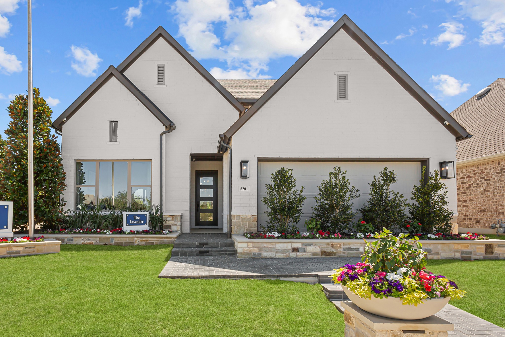 Modern two-story white house with dark trim, large windows, landscaped yard, and a flower planter in the foreground under a partly cloudy sky—showcasing the elegance found in model homes Dallas buyers admire.
