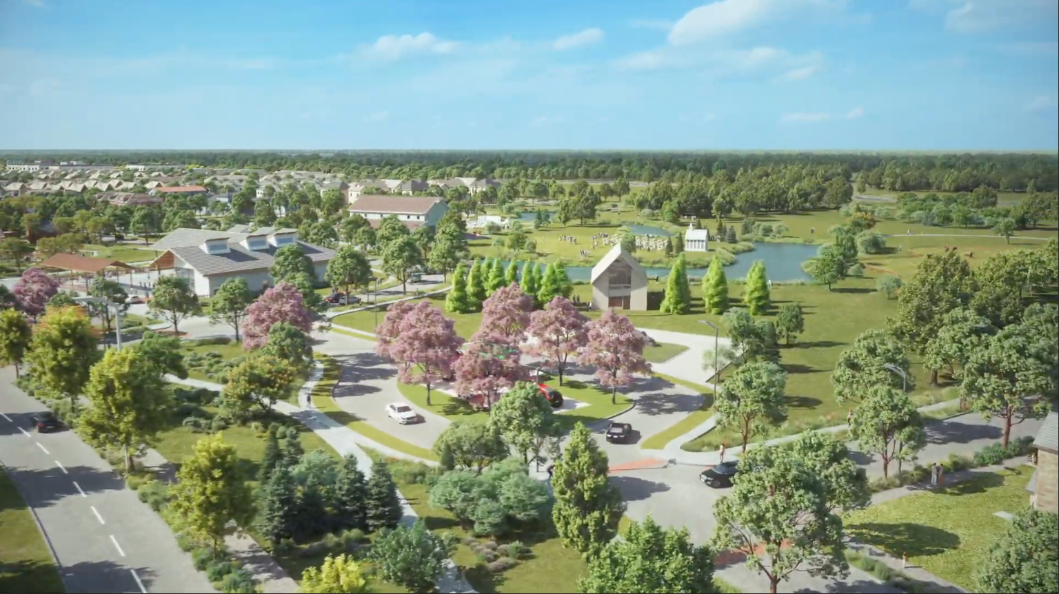 Aerial view of a suburban neighborhood with roads, houses, pink-flowering trees, green lawns, and a pond surrounded by trees under a blue sky.