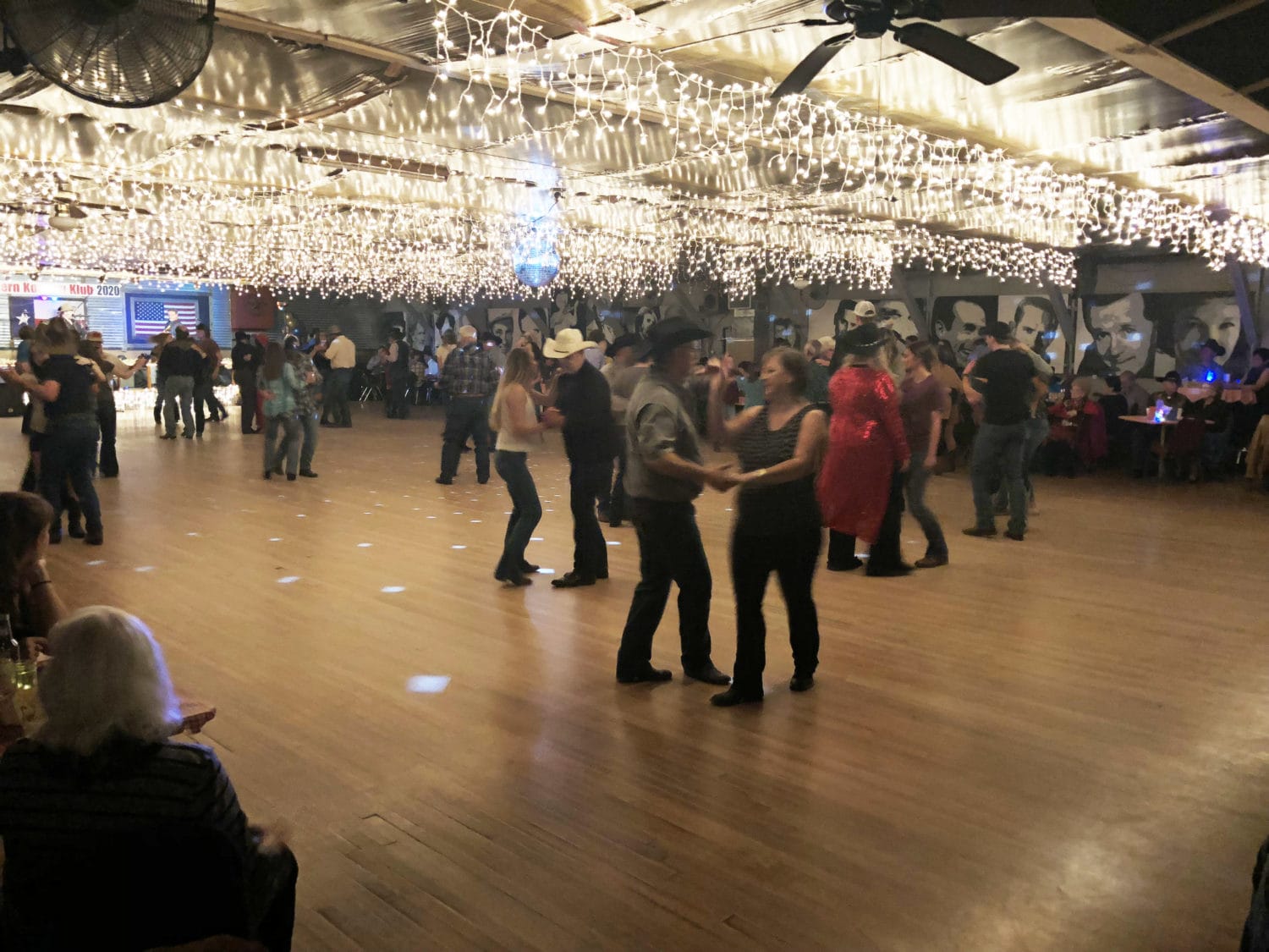People dance in pairs on a wooden floor in a large hall decorated with string lights and mural portraits; some diners sit at tables around the edges.